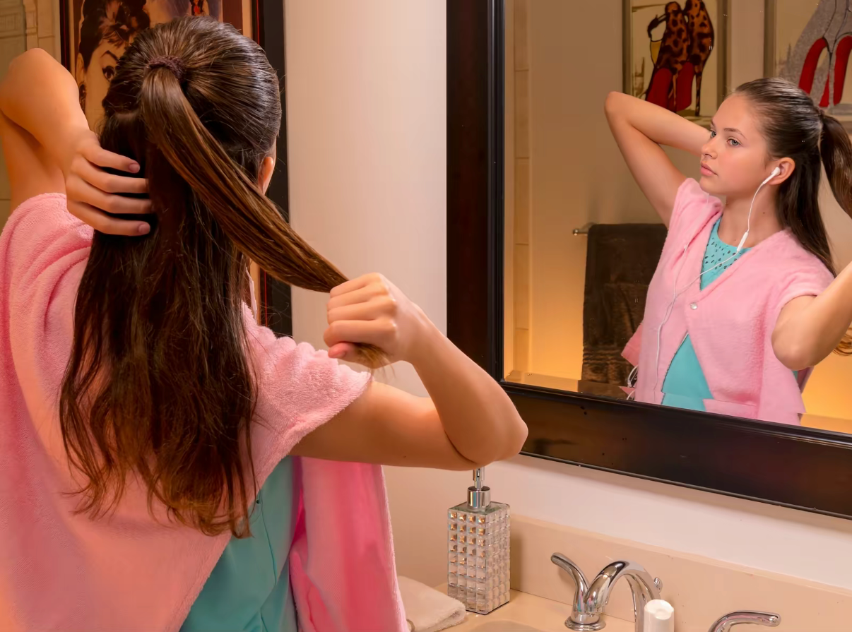 Girl brushing wet hair at the bathroom mirror wearing the pink La Capelette over an aqua top.